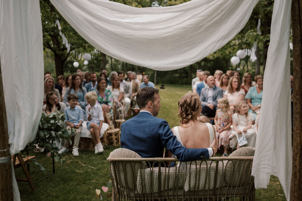 buiten ceremonie vanaf achter gezien, bruidspaar zit samen op bankje en op de achtergrond zie je de gasten